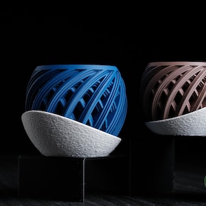 May include: Two decorative spherical planters, one blue and one brown, sit on white textured saucers. The planters have a woven design and are displayed against a black background.