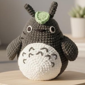 May include: A handmade crochet Totoro plush toy in gray with a white belly and black details. The plush has a green leaf on its head and is displayed on a wooden stand. The toy is approximately 6 inches tall.