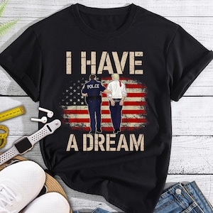 Może przedstawiać: Czarny T-shirt z grafiką przedstawiającą dwie postacie przed amerykańską flagą. Na koszulce nadrukowany jest tekst "I HAVE A DREAM".