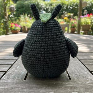 May include: A handmade, dark gray crochet bird with small wings and two upright ears. A small, light green leaf is attached to the top of the bird's head. The bird is sitting on a wooden table outdoors.