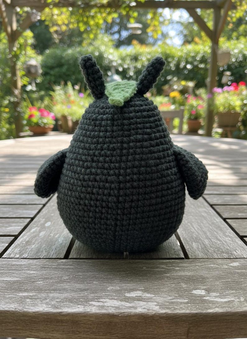 May include: A handmade, dark gray crochet bird with small wings and two upright ears. A small, light green leaf is attached to the top of the bird's head. The bird is sitting on a wooden table outdoors.