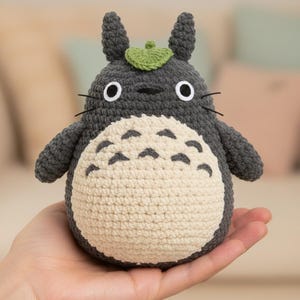 Può includere: Un peluche Totoro all'uncinetto fatto a mano. Il peluche è grigio con una pancia color crema, occhi neri e una foglia verde sulla testa. Il Totoro è tenuto nella mano di una persona.