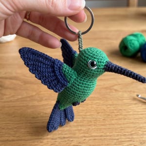 Puede incluir: Un llavero de colibrí tejido a ganchillo con cuerpo verde, alas azul oscuro y cola azul oscuro. El pájaro tiene un pico largo y puntiagudo de color azul oscuro y un ojo de plástico negro y blanco visible. Un anillo de metal plateado está sujeto en la parte superior.