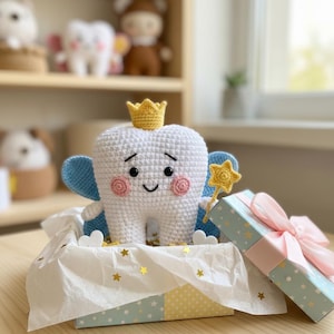May include: A crocheted tooth fairy doll with a gold crown, blue wings, and a star wand, presented in a gift box with a pink ribbon. The tooth has rosy cheeks and a smiling face. Other crocheted toys are in the background.