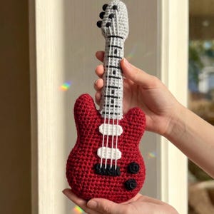 Op de afbeelding: Een gehaakte rode elektrische gitaar met een grijze hals en kop. De gitaar heeft witte en zwarte details, waaronder snaren en knoppen. Het handgemaakte instrument wordt in de handen van een persoon gehouden.