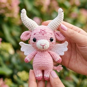 Könnte beinhalten: Eine handgefertigte rosa Häkelziege mit weißen Hörnern, Flügeln und einem Schwanz. Die Ziege hat schwarze Augen und ein Pentagramm auf der Stirn. Die Ziege wird von einer Person gehalten.