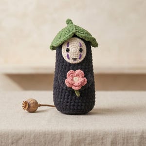 Puede incluir: Muñeca de ganchillo con forma de personaje de El Viaje de Chihiro, con cuerpo gris oscuro, cara blanca y sombrero en forma de hoja verde. La muñeca sostiene una flor rosa y tiene marcas moradas. Una cápsula de semilla seca descansa cerca.
