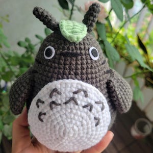 May include: Handmade crochet plush toy in the shape of a Totoro character. The plush is primarily dark gray with a white belly and black markings. It has large, round eyes and small ears with a green leaf on top.