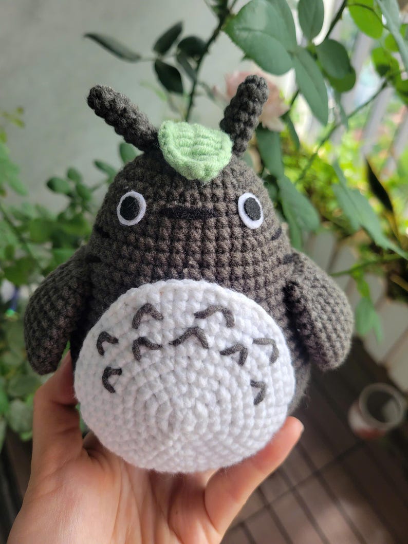 May include: Handmade crochet plush toy in the shape of a Totoro character. The plush is primarily dark gray with a white belly and black markings. It has large, round eyes and small ears with a green leaf on top.