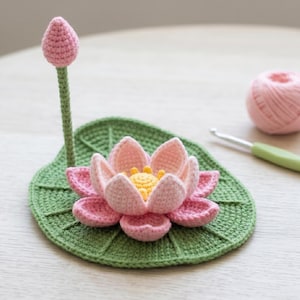 Puede incluir: Un conjunto de flor de loto tejida a ganchillo que incluye una base grande de hoja de nenúfar verde, un loto rosa en flor con centro amarillo y un capullo de loto rosa sobre un tallo verde. Un ovillo de lana rosa y un ganchillo están cerca.