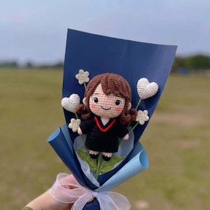 May include: A handmade crochet doll in a graduation gown, holding a bouquet of white heart and star-shaped flowers. The doll has brown hair and is wrapped in blue paper, tied with a white ribbon. The background is blurred.