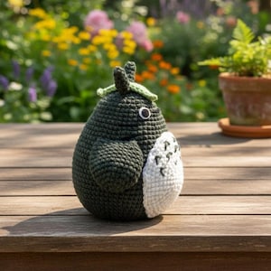May include: A handmade, dark green crochet figure with a white belly patch and a leaf-shaped hat. The figure has a single black eye and is sitting on a wooden surface. The background features a blurred garden with colorful flowers and a potted plant.
