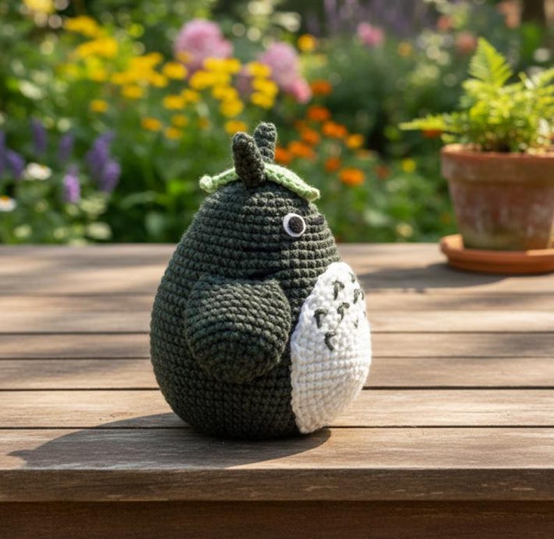 May include: A handmade, dark green crochet figure with a white belly patch and a leaf-shaped hat. The figure has a single black eye and is sitting on a wooden surface. The background features a blurred garden with colorful flowers and a potted plant.