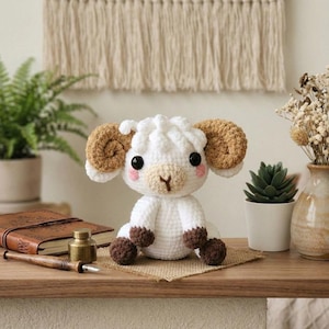 May include: A handmade white crochet ram with brown horns, hooves, and a tan face. The amigurumi animal has black eyes and pink cheeks. It sits on a small burlap square on a wooden shelf, with a fern, succulent, and decorative items in the background.
