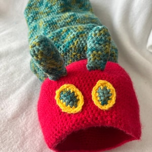 Puede incluir: Un gorro de oruga hecho a mano con ganchillo en colores brillantes. El gorro presenta una cabeza roja con ojos con borde amarillo y un cuerpo turquesa y amarillo. El gorro está diseñado para ser usado en la cabeza.