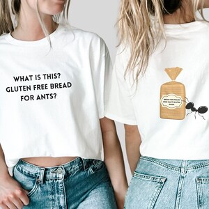 Puede incluir: Camiseta blanca recortada con el texto "What is this? Gluten free bread for ants?" en la parte delantera. La parte posterior de la camiseta presenta una hormiga de dibujos animados que lleva una bolsa de pan etiquetada como "Bread for folks who can't gluten food."