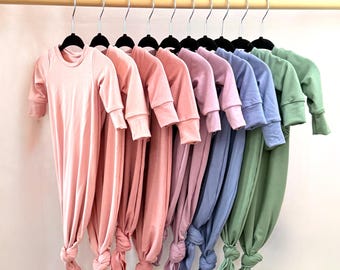May include: A row of eight knotted baby gowns in various pastel colors, including pink, lavender, blue, and green, hanging on a wooden rack.