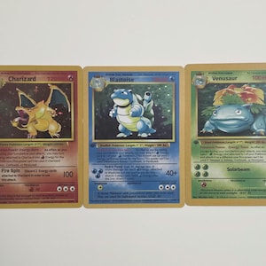 May include: Three rectangular trading cards featuring Charizard, Blastoise, and Venusaur. The red Charizard card, blue Blastoise card, and green Venusaur card each display character illustrations, text, and symbols on a gold-bordered background.