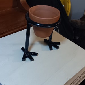 May include: A black metal plant stand with three legs shaped like bird feet. A terracotta pot sits on top of the stand.