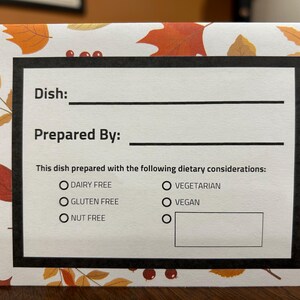May include: A white card with a black border, featuring text fields for "Dish:" and "Prepared By:". It includes checkboxes for dietary considerations: Dairy Free, Gluten Free, Nut Free, Vegetarian, and Vegan. The card has a fall-themed leaf and berry pattern.