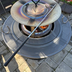 May include: A round, gray metal fire pit with a perforated surface and a lid. The lid is open, revealing a fire inside. A long, black-handled poker rests on the side. Horseshoe-shaped hooks are attached to the side of the fire pit.