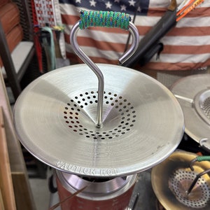 May include: A silver camping stove with a metal lid and a warning label that reads "CAUTION HOT". The stove has a green and red rope handle.