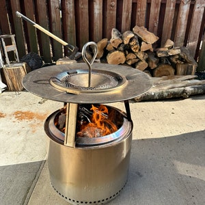 Pu&ograve; includere: Un focolare Solo Stove Bonfire in acciaio inossidabile con un fuoco a legna all'interno. Il focolare ha una parte superiore rotonda e piatta con un manico e una base rotonda e cilindrica con un logo. Il focolare &egrave; posizionato su un patio in cemento.