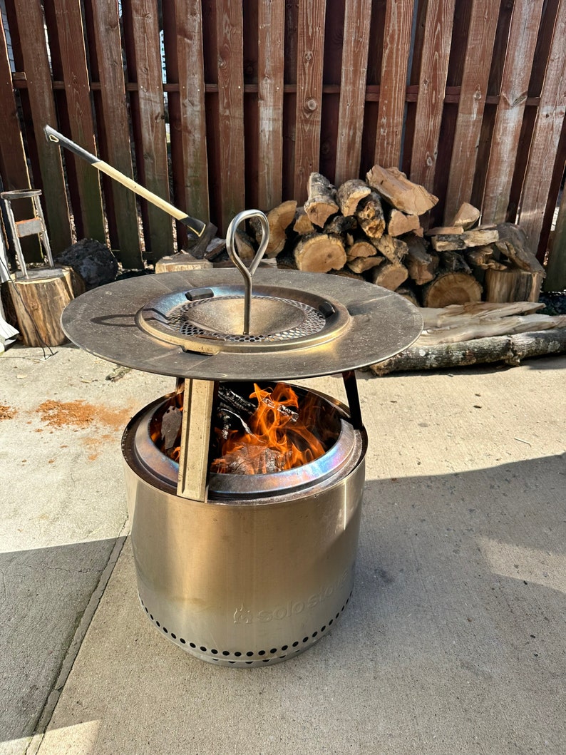 Pu&ograve; includere: Un focolare Solo Stove Bonfire in acciaio inossidabile con un fuoco a legna all'interno. Il focolare ha una parte superiore rotonda e piatta con un manico e una base rotonda e cilindrica con un logo. Il focolare &egrave; posizionato su un patio in cemento.