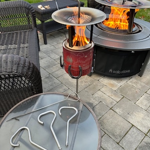 May include: A Solo Stove bonfire pit with a red and black design, a metal fire grate, and a metal lid. The fire pit is lit and has flames coming out of the top. There are three metal hooks on the side of the fire pit. The fire pit is sitting on a patio with a glass table and a wicker chair in the background.