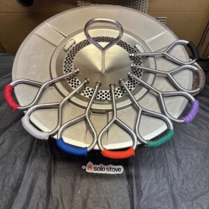 Pu&ograve; includere: Una stufa Solo Stove argentata con una base rotonda e una maniglia al centro. Ci sono otto maniglie metalliche con cordini di diversi colori avvolti attorno a loro. I cordini sono rosso, blu, verde, viola, grigio e nero.
