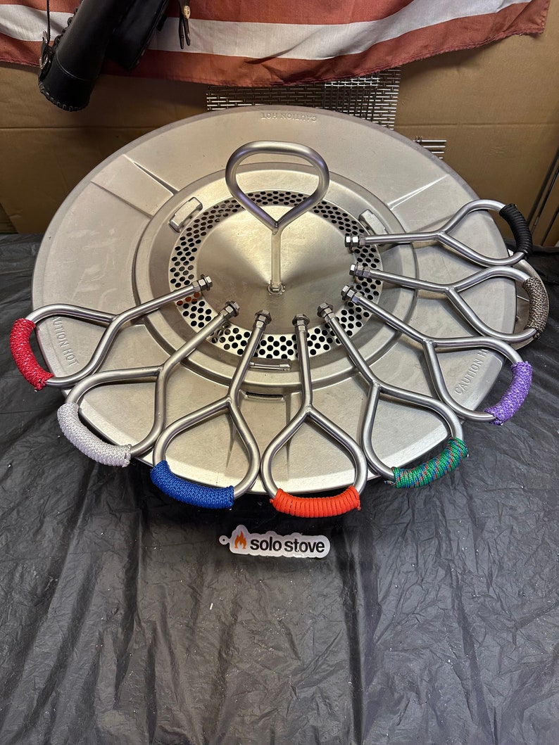 Pu&ograve; includere: Una stufa Solo Stove argentata con una base rotonda e una maniglia al centro. Ci sono otto maniglie metalliche con cordini di diversi colori avvolti attorno a loro. I cordini sono rosso, blu, verde, viola, grigio e nero.