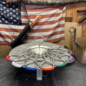 Pu&ograve; includere: Una stufa Solo Stove in acciaio inossidabile con una custodia di trasporto nera e un logo che dice "solo stove". La stufa ha un anello di manici in metallo con avvolgimenti in paracord colorati. La stufa &egrave; appoggiata su una superficie nera.