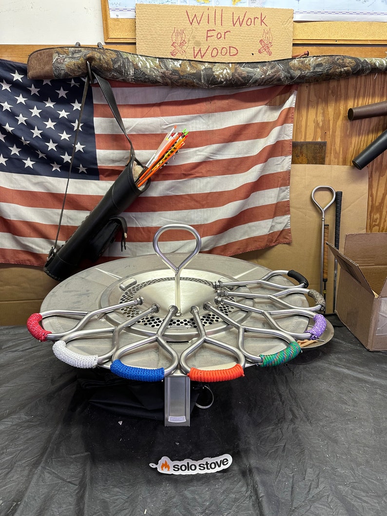 Pu&ograve; includere: Una stufa Solo Stove in acciaio inossidabile con una custodia di trasporto nera e un logo che dice "solo stove". La stufa ha un anello di manici in metallo con avvolgimenti in paracord colorati. La stufa &egrave; appoggiata su una superficie nera.