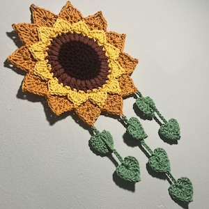Puede incluir: Un tapiz de pared de girasol tejido a crochet con un centro marrón y pétalos amarillos. El girasol tiene hojas verdes que cuelgan de la parte inferior.