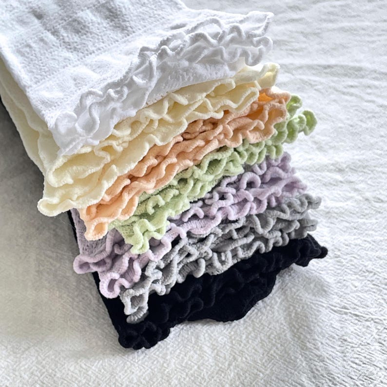 May include: A stack of ruffled socks in various pastel colors, including white, yellow, peach, green, lavender, gray, and black. The socks have a soft, textured appearance and a delicate, feminine design.