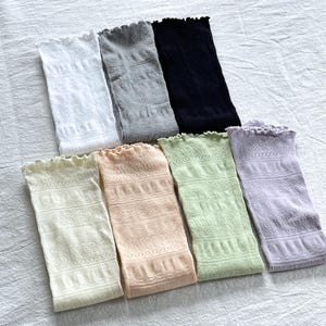 May include: An assortment of seven open-weave, ruffled-top arm sleeves in various colors. The colors include white, gray, black, cream, peach, light green, and lavender. The sleeves have a delicate, lacy pattern.