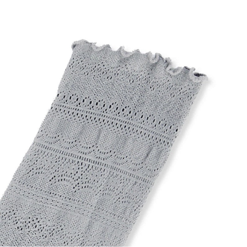 May include: A light gray, open-weave textile with a scalloped edge. The fabric has a delicate, lacy pattern with various geometric shapes and lines. The textile is likely a sleeve or part of a garment.