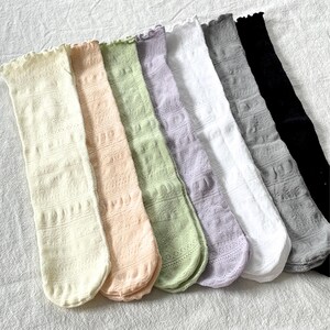 May include: A collection of seven knee-high socks in various pastel colors, including cream, peach, light green, lavender, white, gray, and black. Each sock features a delicate, openwork pattern and a ruffled top edge.