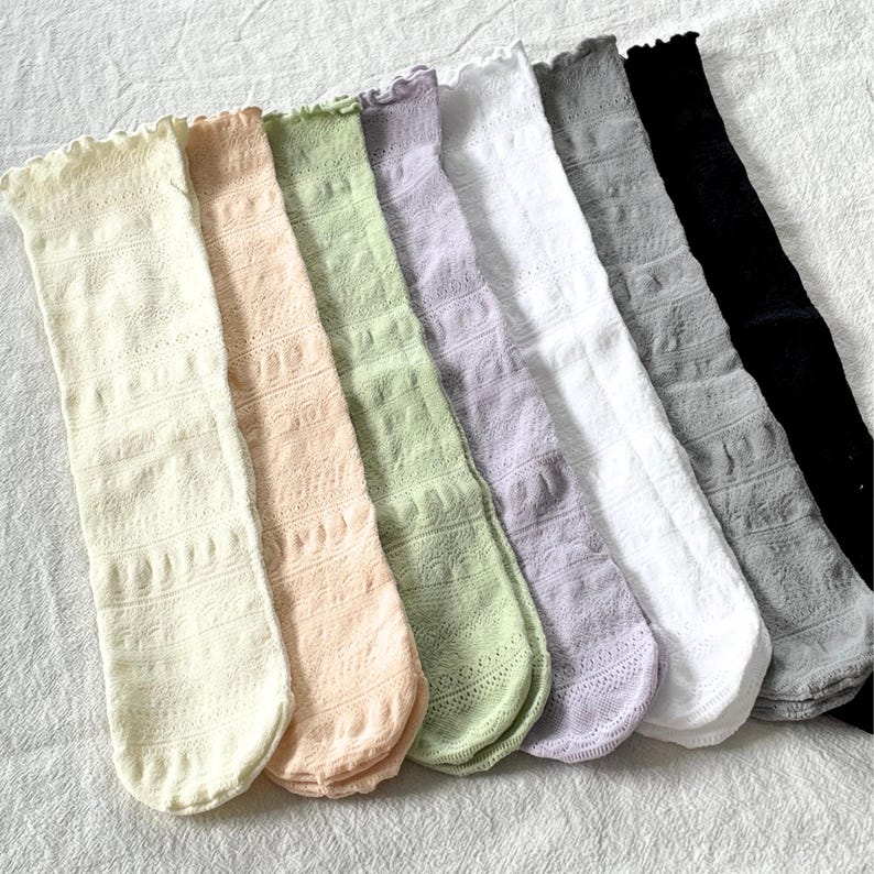 May include: A collection of seven knee-high socks in various pastel colors, including cream, peach, light green, lavender, white, gray, and black. Each sock features a delicate, openwork pattern and a ruffled top edge.