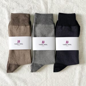 May include: Three pairs of socks in brown, gray, and black, each with a herringbone pattern. The socks have a contrasting color at the toe and cuff. Each pair is wrapped with a white band featuring the text "YANG MAL" and a logo.