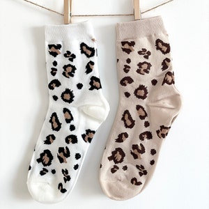May include: A pair of socks with a leopard print design. One sock is white with brown spots, and the other is beige with brown spots.