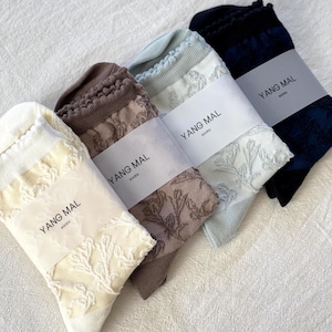 Puede incluir: Un surtido de calcetines en crema, marrón, azul claro y marino. Cada par está envuelto con una etiqueta que dice "YANG MAL socks". Los calcetines presentan un delicado diseño floral.
