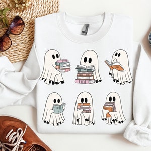 May include: White sweatshirt with a graphic of six ghosts reading books. The ghosts are white with black eyes and mouths. The books are colorful and stacked. The sweatshirt is a light grey color.