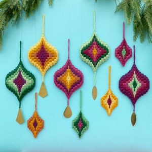 May include: Nine colourful, geometric, embroidered ornaments hang from a string. The ornaments are in shades of green, yellow, red, and purple. Each ornament has a small, gold, metal bead at the bottom.