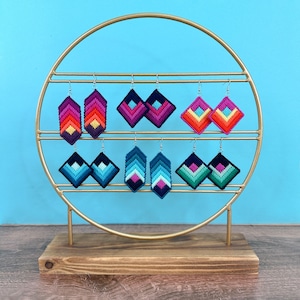 May include: A gold metal stand with a round top and three legs displays eight pairs of colorful geometric patterned earrings. The earrings are made of yarn and are in various shades of blue, pink, red, and yellow.