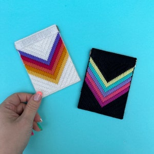May include: Two needlepoint cardholders. One is white with a rainbow chevron design, the other is black with a pastel chevron pattern. Both have a silver metal clasp. The cardholders are on a light blue background.