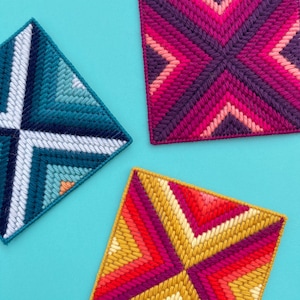 Może przedstawiać: Trzy kwadratowe, haftowane ścienne wiszące z geometrycznymi wzorami. Górne wiszące jest fioletowe i różowe, lewe wiszące jest niebieskie i białe, a dolne wiszące jest żółte, czerwone i różowe.