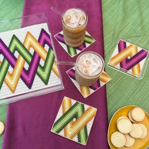Puede incluir: Una bandeja y posavasos de acrílico transparente con un diseño geométrico en verde, amarillo y morado. Dos bebidas de café helado con pajitas se encuentran sobre los posavasos. Un plato amarillo contiene galletas.