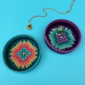Puede incluir: Dos cajas redondas para joyas de punto de aguja bordadas y coloridas. Una caja es turquesa con un diseño de estrella estrellada rosa, naranja y amarilla. La otra caja es morada con un diseño de estrella estrellada verde, morado y azul. Dos anillos dorados están dentro de la caja morada.