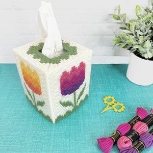 May include: A white tissue box cover with a colorful floral design. The cover features two tulips, one orange and yellow and the other pink and purple. The cover is made of plastic canvas and is decorated with embroidery floss.